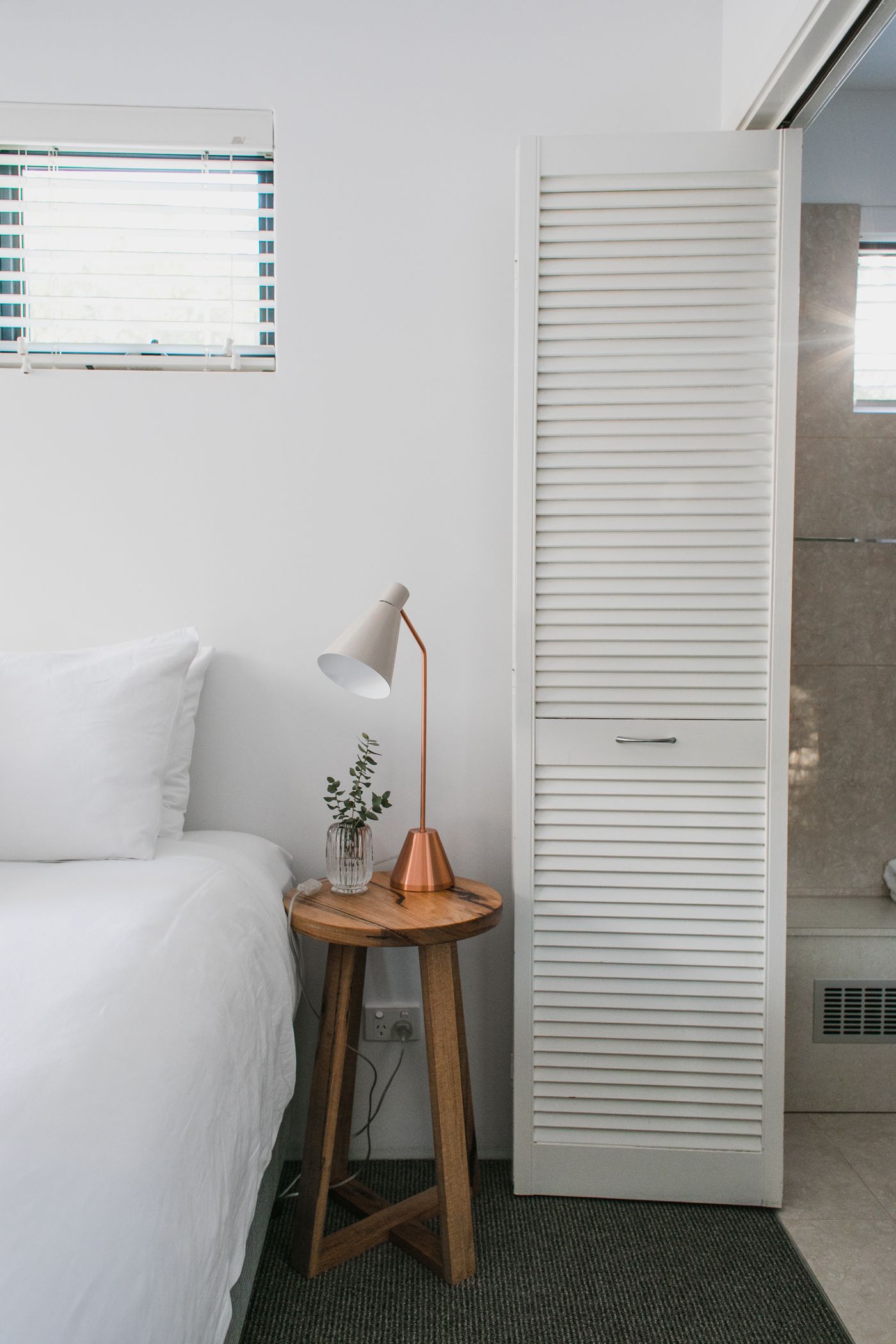 A small wooden bedside table holding a reading lamp and flowers.