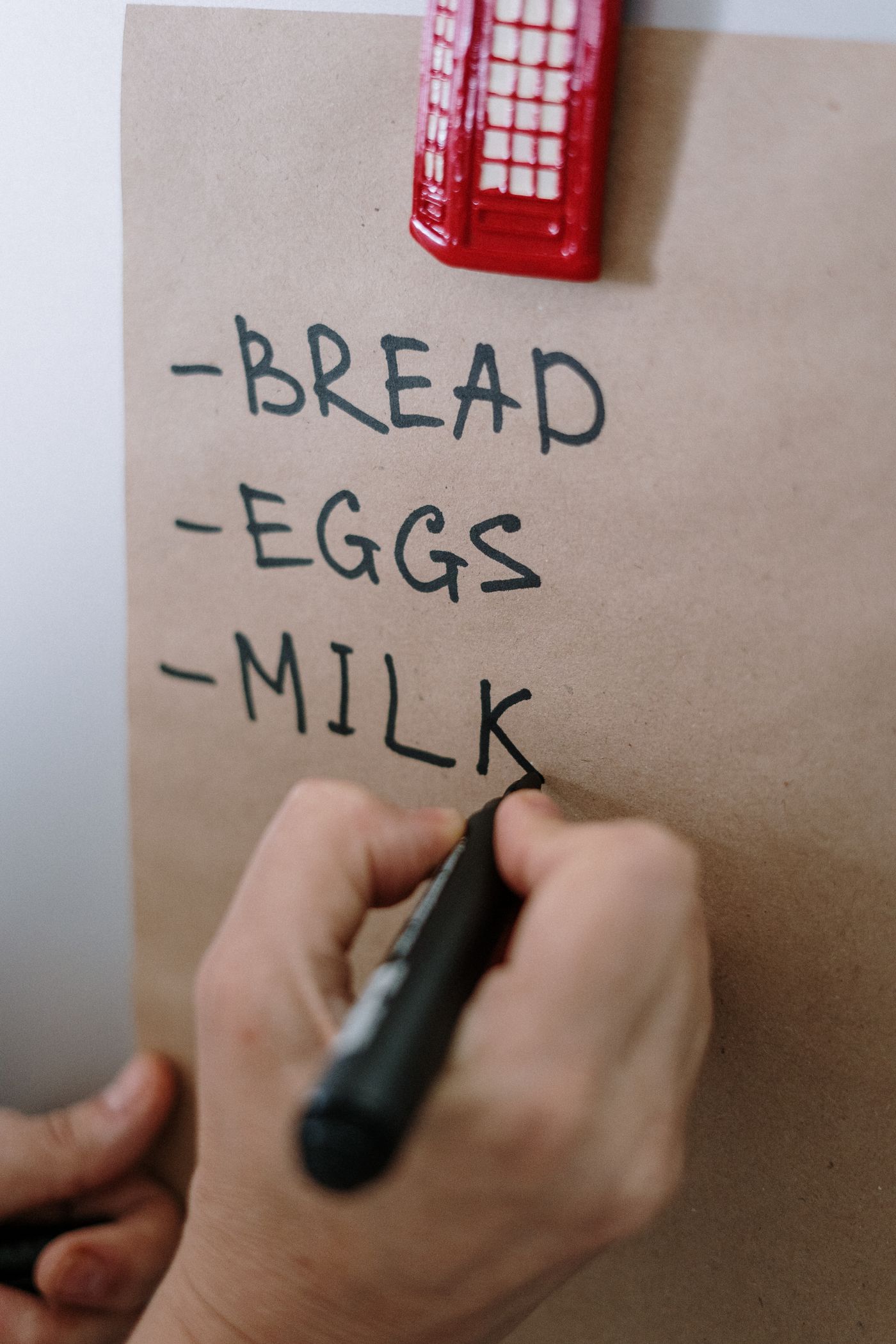 A hand writing bread, eggs and milk on a paper shopping list.