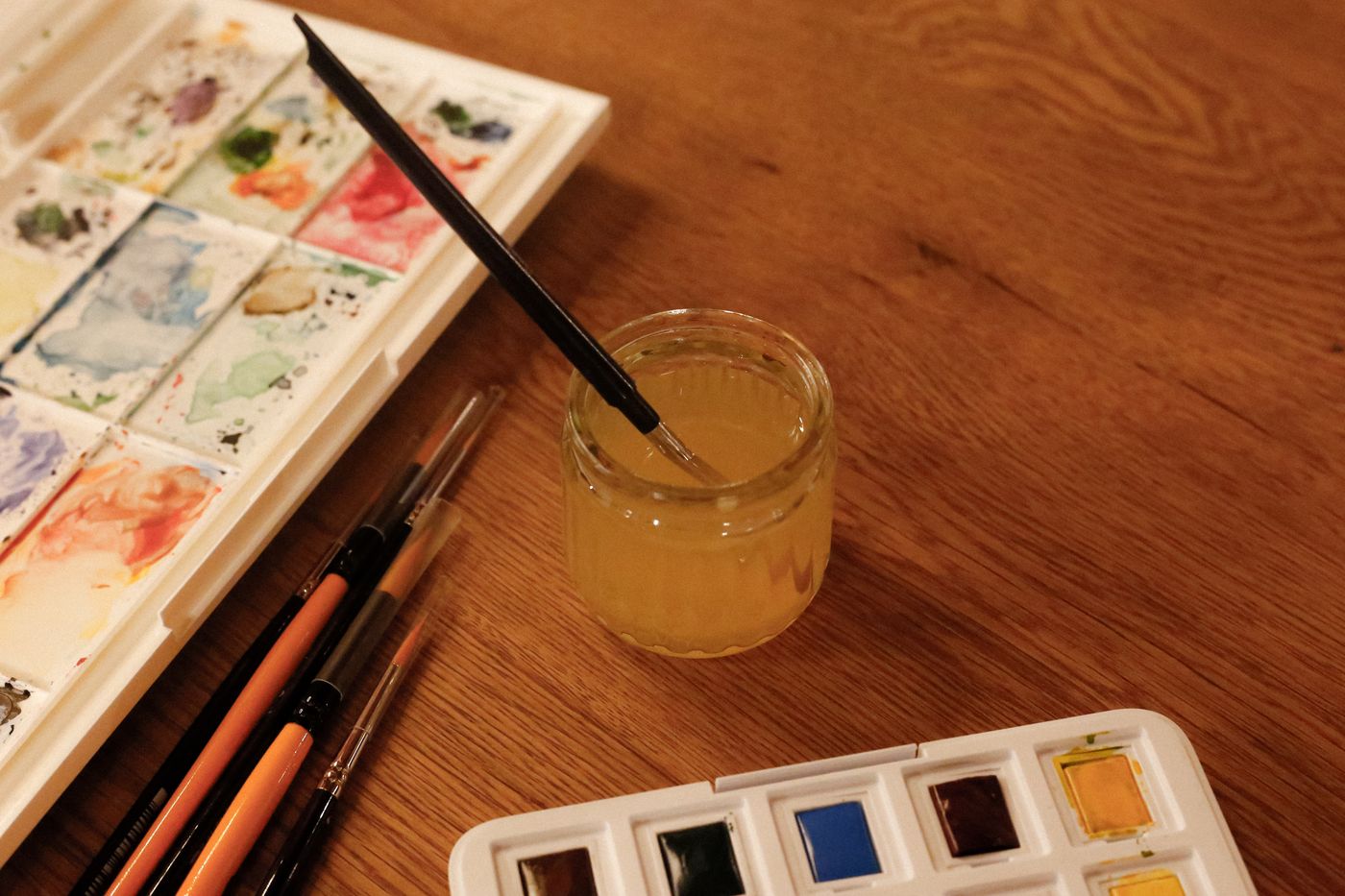 Watercolour paints, brushes and a water jar on a wooden table.